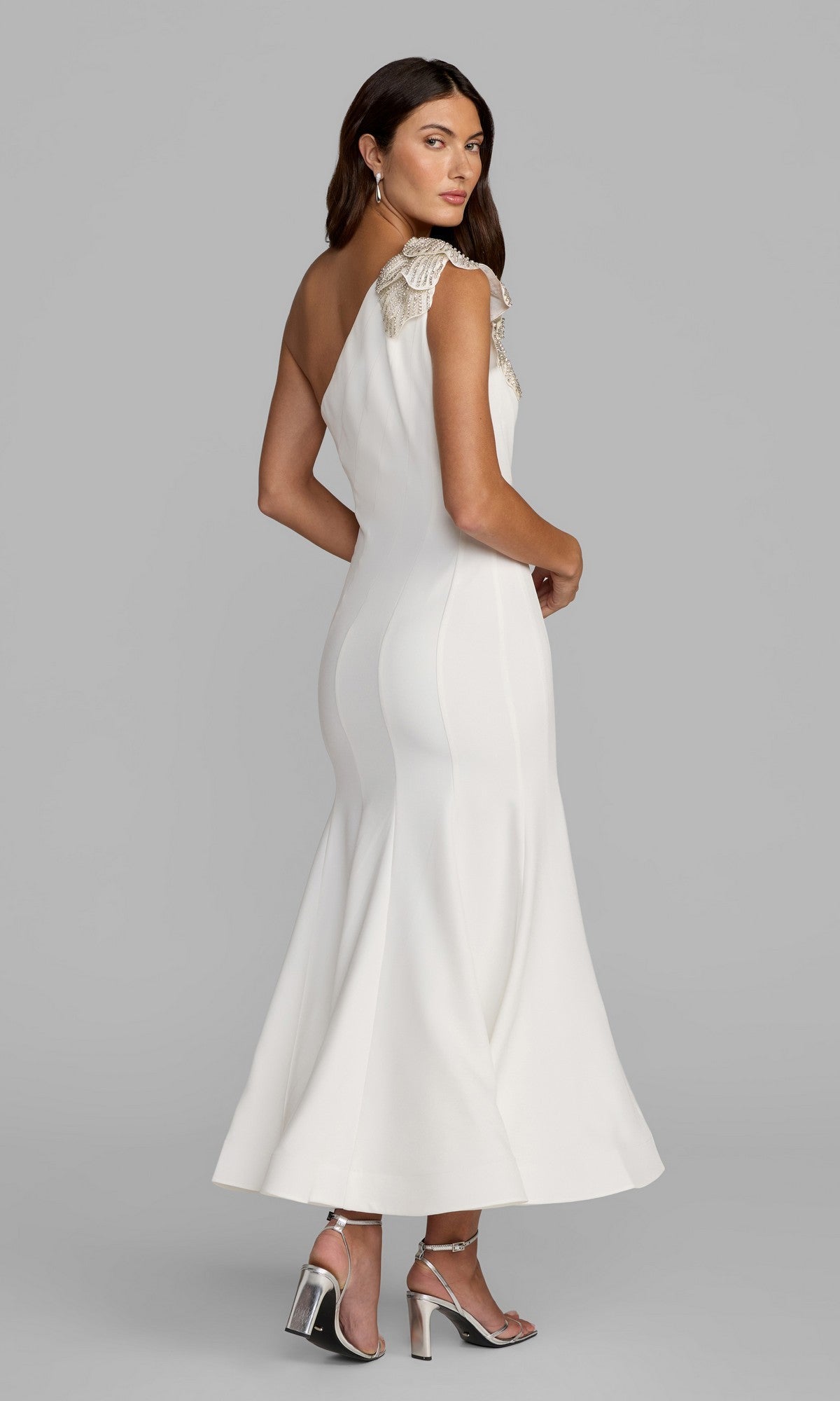 A woman in the Tea-Length Party Dress: Mac Duggal 13221, a fitted crepe one-shoulder midi dress with an embellished floral shoulder applique, paired with silver high-heeled sandals, stands against a plain gray background.